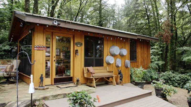 Wooden cabin with a bench on a porch, surrounded by lush greenery. Warm lighting highlights the rustic charm and inviting outdoor space.