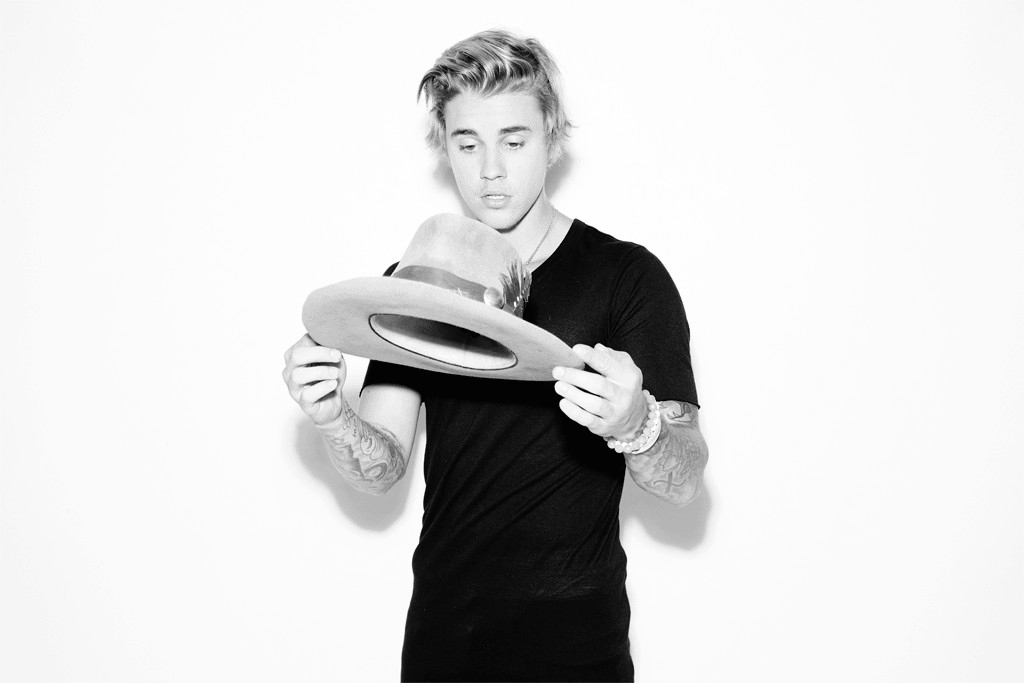 Studio portrait of a young adult with blonde hair holding a tan hat with a black shirt against a bright white background.