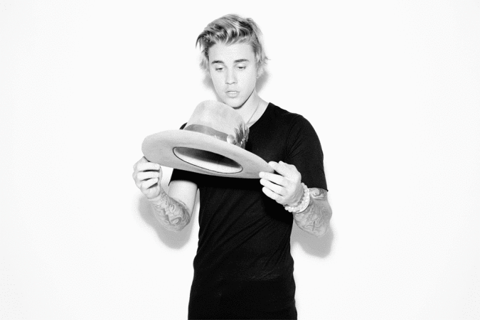 Studio portrait of a young adult with blonde hair holding a tan hat with a black shirt against a bright white background.