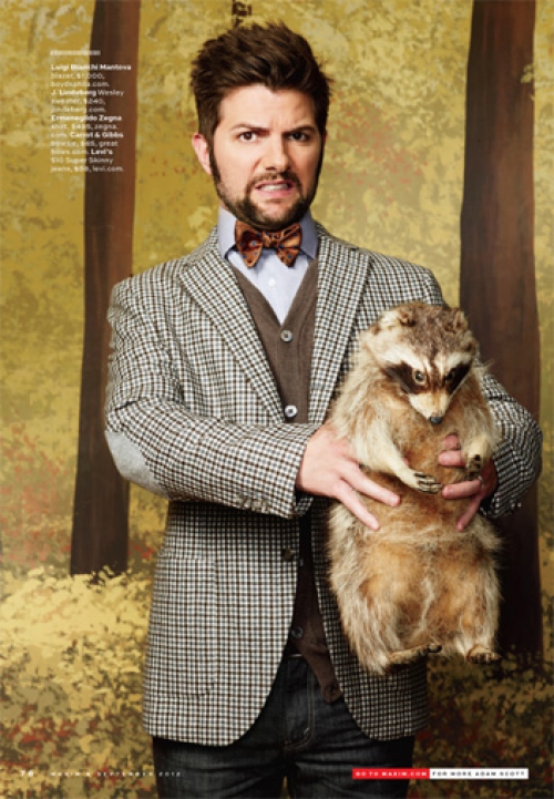 Adult male with dark hair and beard wearing a blazer and bow tie, holding a stuffed raccoon in front of a forest background.