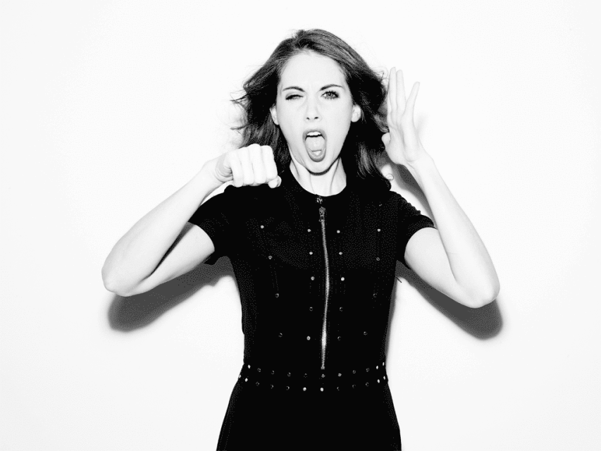 A medium shot captures a young woman with dark hair and a black dress gesturing with her hands and screaming against a white backdrop.