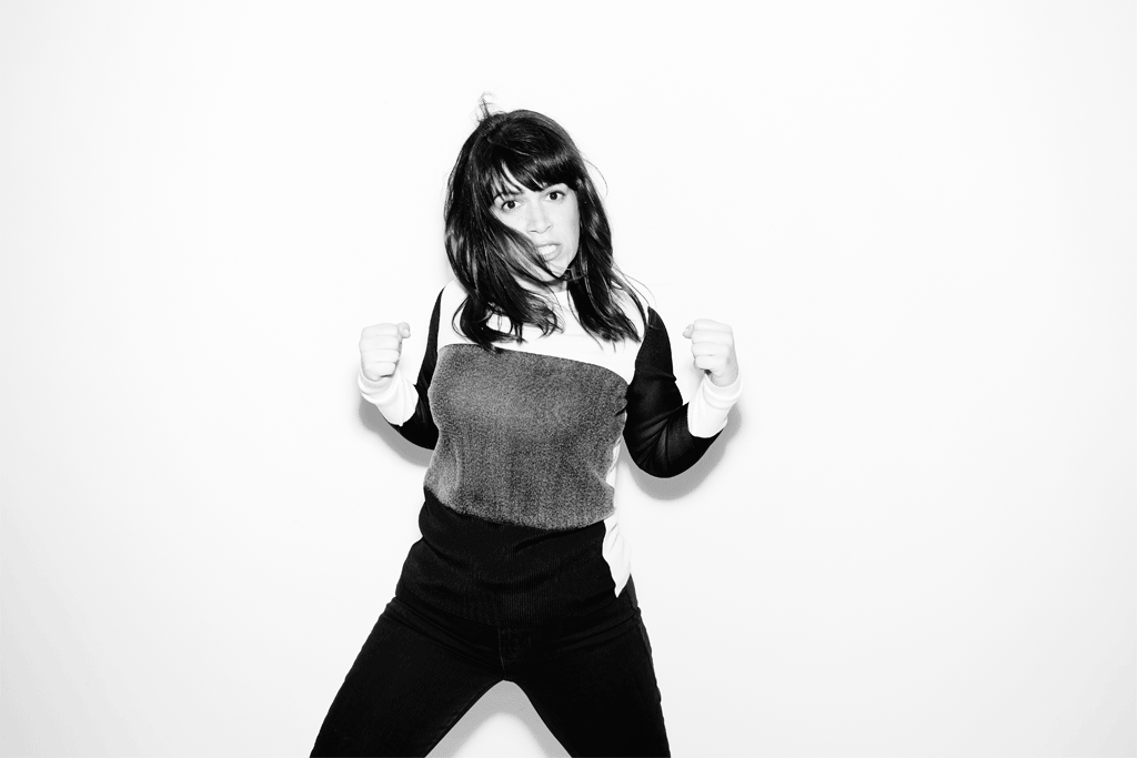 Black and white portrait of a young adult woman with dark hair wearing a two-tone sweater, raising her fists against a white backdrop.