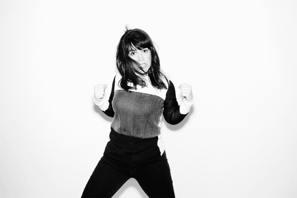 Black and white portrait of a young adult woman with dark hair wearing a two-tone sweater, raising her fists against a white backdrop.