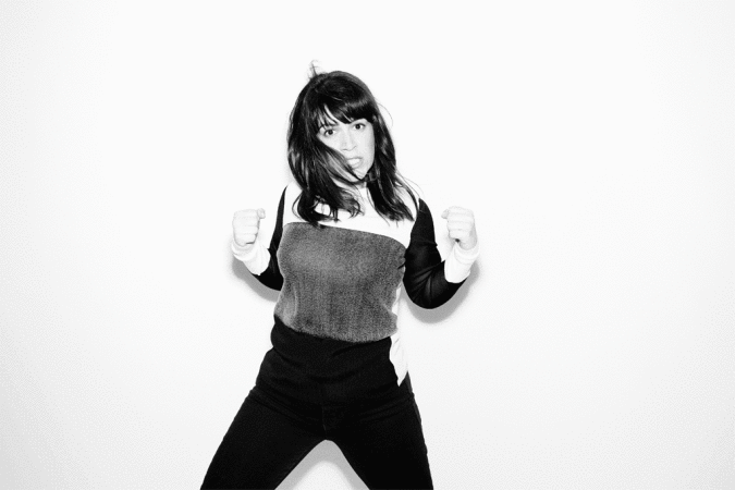 Black and white portrait of a young adult woman with dark hair wearing a two-tone sweater, raising her fists against a white backdrop.