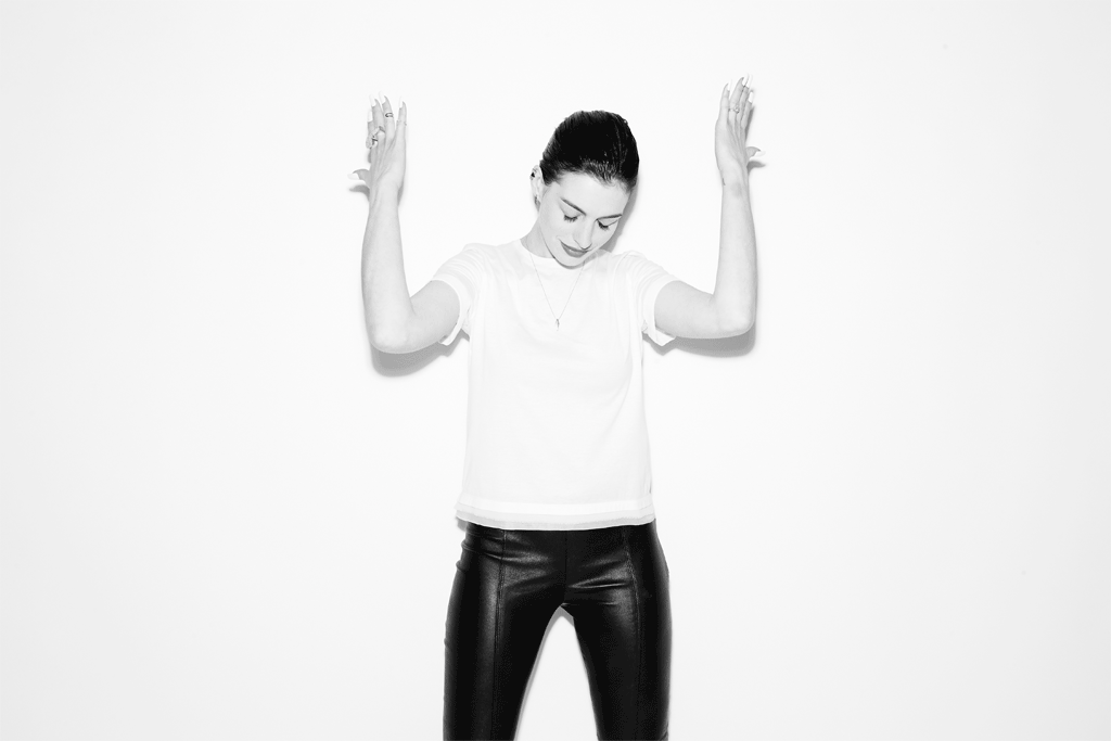 Anne Hathaway in a white t-shirt and black pants posing with hands raised against a white background by Peter Yang
