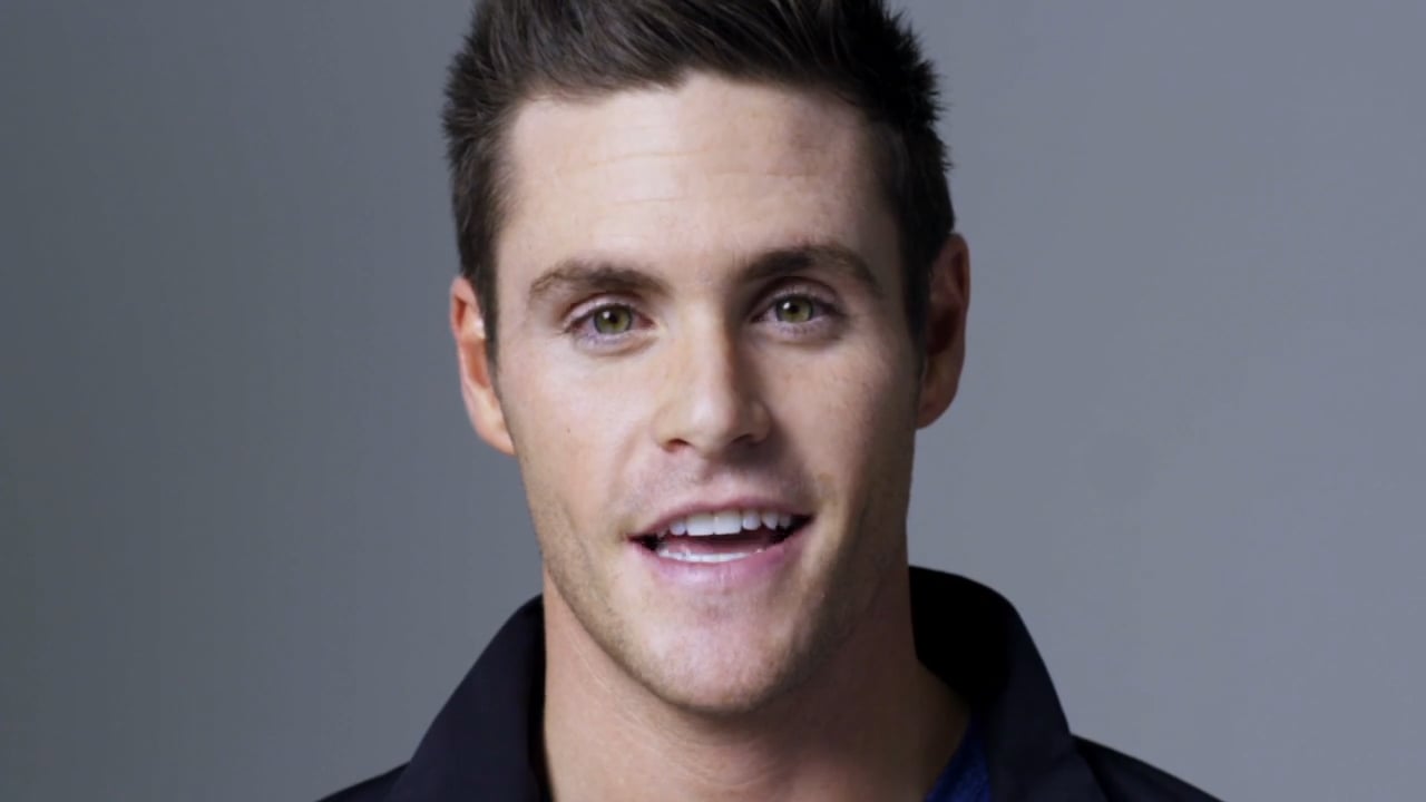 Close-up portrait of a man in his 30s smiling against a neutral grey background.