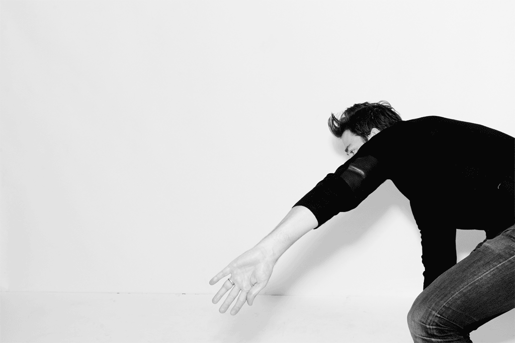 Monochrome portrait of an adult male reaching out against a white wall, wearing a dark long-sleeve shirt and denim jeans.