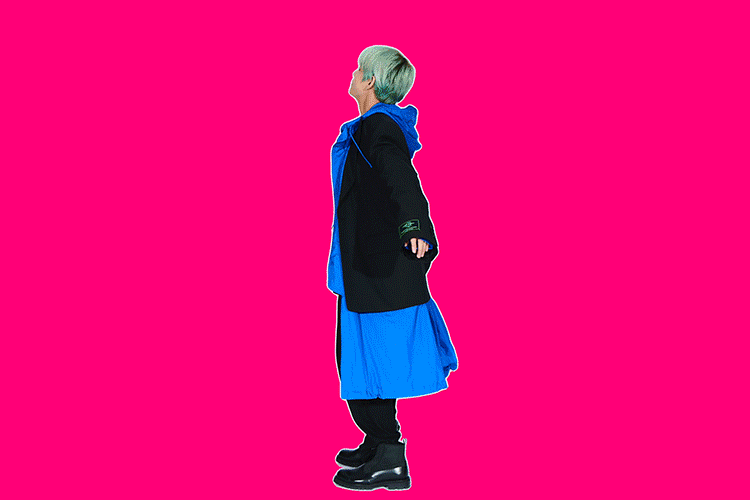 Woman in black coat and blue dress standing on a pink background. She has short grey hair and is wearing black boots.