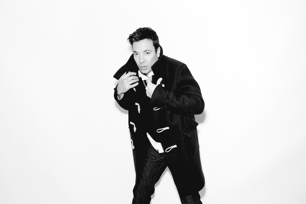 Black and white portrait of a man in a duffle coat standing against a white backdrop, looking at the camera with a neutral expression.