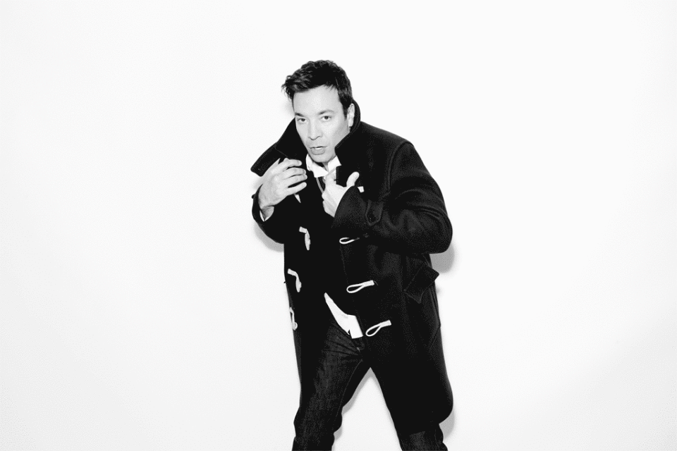 Black and white portrait of a man in a duffle coat standing against a white backdrop, looking at the camera with a neutral expression.