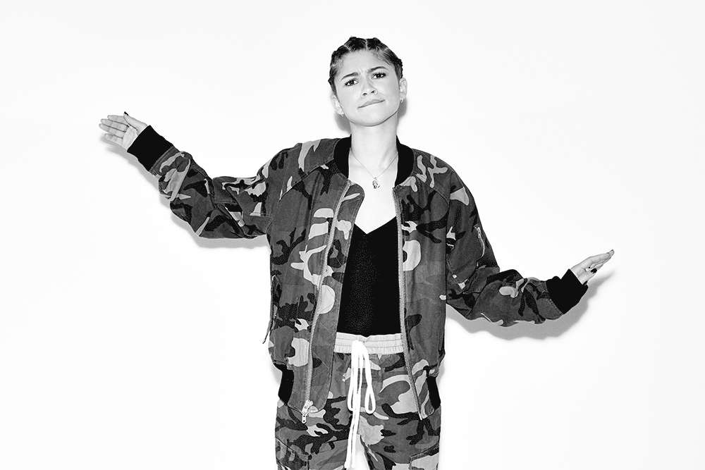 Black-and-white portrait of a female adult wearing a camouflage jacket and pants, with her arms extended to both sides against a white background.