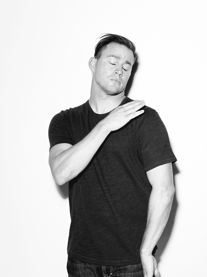 Black and white portrait of a man with his eyes closed, wearing a dark t-shirt and jeans, posing in front of a white wall.