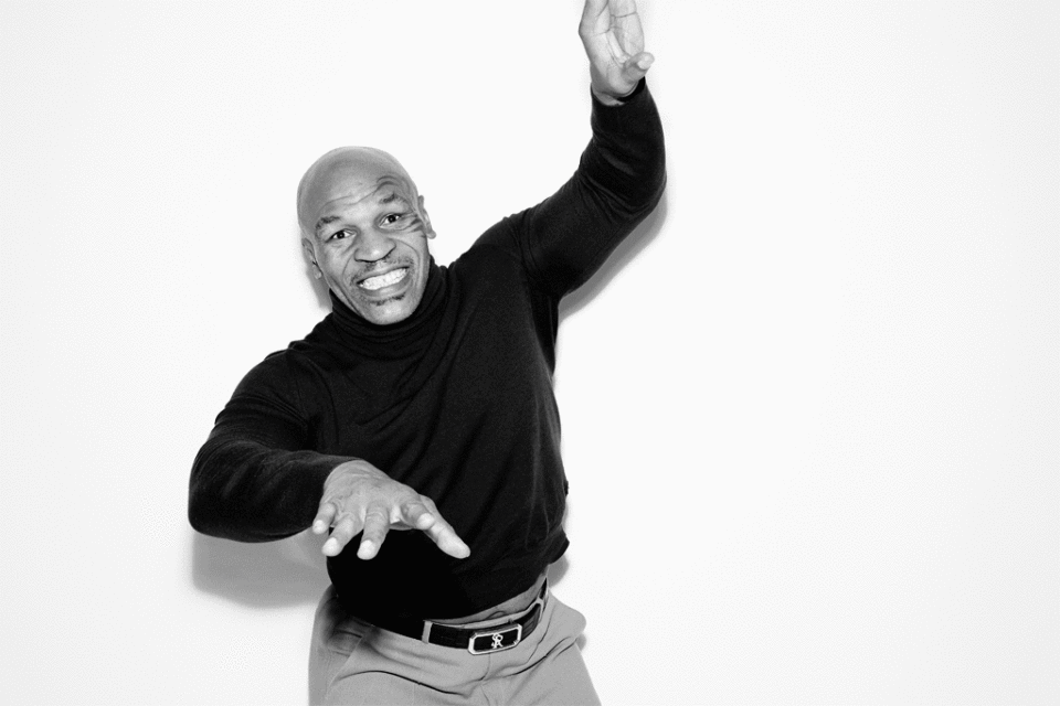 Black and white portrait of Mike Tyson in turtleneck and slacks gesturing with his hands indoors against a white wall backdrop.