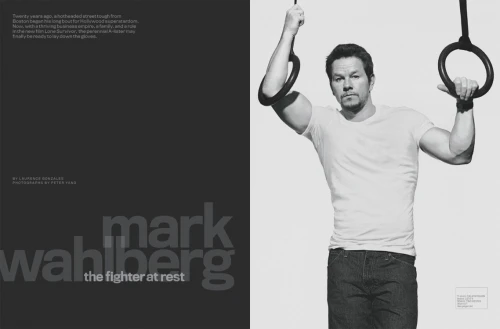 Black and white portrait of Mark Wahlberg, wearing a white t-shirt and blue jeans, suspended from gymnastic rings against a plain white background.
