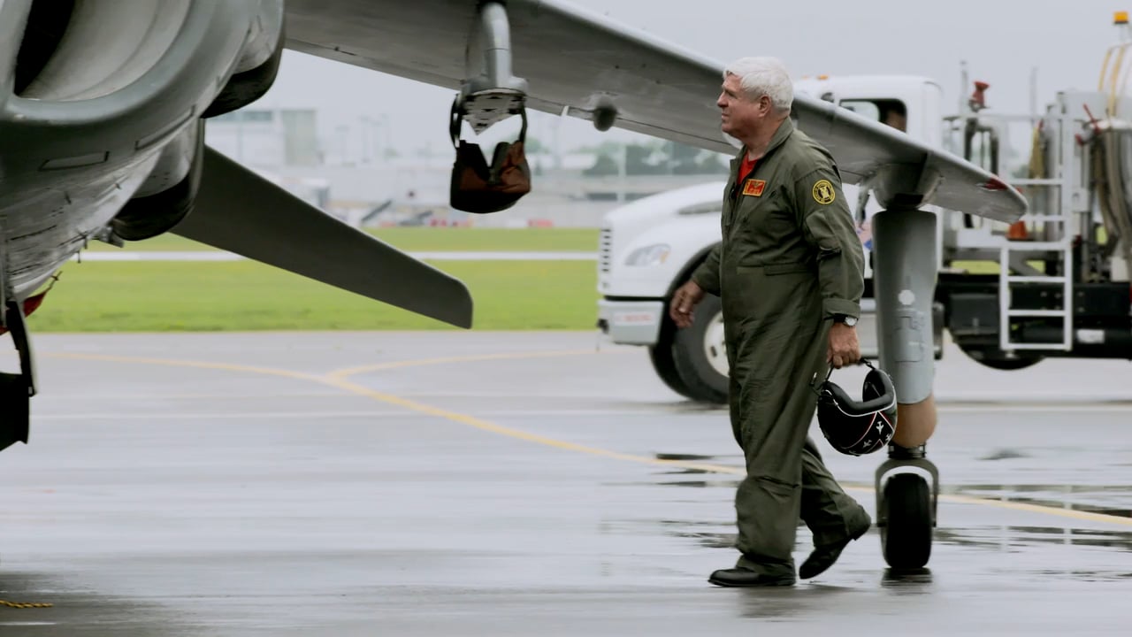 An adult male walks on an airport tarmac near an airplane with a refueling truck in the background on a cloudy day.