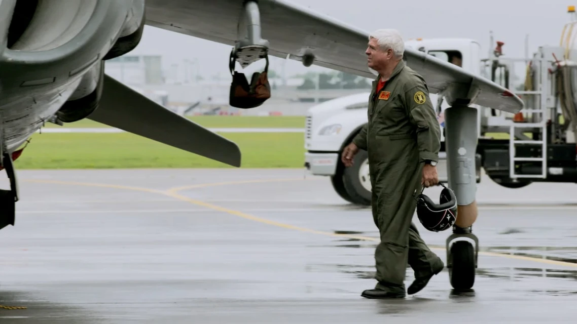 An adult male walks on an airport tarmac near an airplane with a refueling truck in the background on a cloudy day.