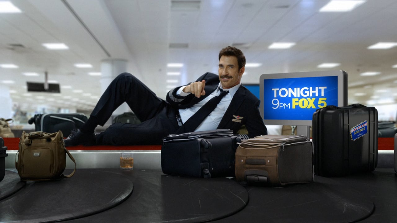 Man in pilot uniform reclining on luggage carousel, pointing forward in airport. Luggage and TV screen in background. Travel concept.