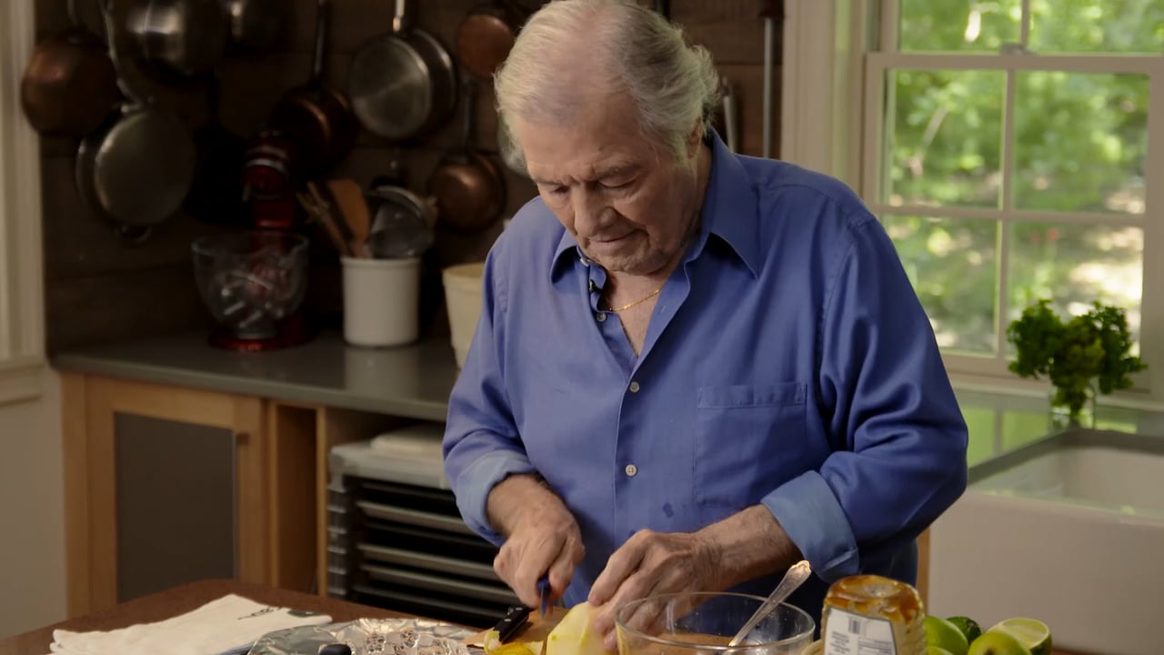 Elderly male chef with white hair cuts fruit on a counter in a kitchen with copper pans hanging on the wall, indoor shot.