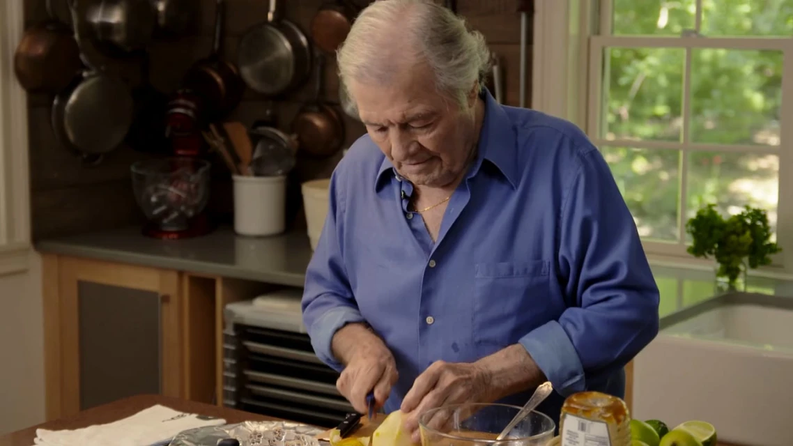 Elderly male chef with white hair cuts fruit on a counter in a kitchen with copper pans hanging on the wall, indoor shot.