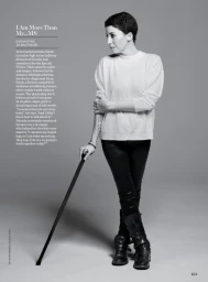 A woman with short dark hair wearing a white sweater and dark trousers leaning on a cane against a grey studio backdrop.