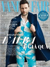 Magazine cover portrait of a man with a beard wearing a blue cloud-print suit and a dark blue coat standing before a brick wall.