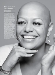 Portrait of a smiling bald woman resting her chin on her hands, wearing earrings and a light top against a grey background.