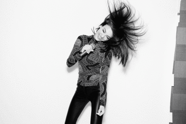 A smiling woman singing into a microphone with dark hair whipping through the air, wearing a patterned top and dark trousers.
