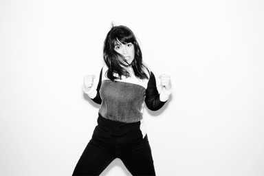 Black and white portrait of a woman with dark hair striking a playful fighting pose against a plain white background.