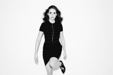 Alison Brie in a black dress with studded trim and dark shoes against a plain white background