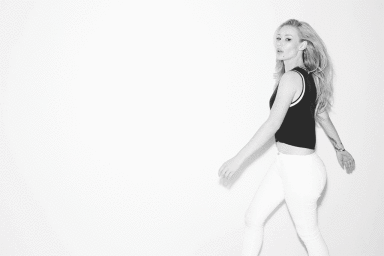 Iggy Azalea wearing a black sleeveless top and white jeans posing against a plain white background in a portrait by Peter Yang.