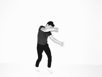 Man wearing a dark t-shirt, dark trousers, and white sneakers posing dynamically against a white background.