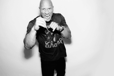 Dwayne Johnson raising his fists in a fighting stance, wearing a t-shirt against a plain white background.