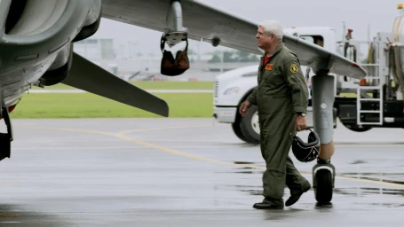 An older man in a green flight suit walking on an airfield holding a helmet with an aircraft wing overhead.