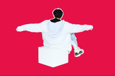 Person seen from behind wearing an oversized white hoodie, sitting on a white cube against a vibrant pink background.