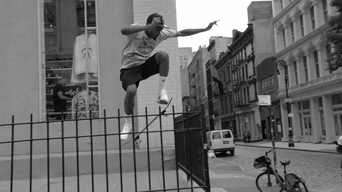 Black and white shot of a skateboarder performing a trick on a rail in an urban setting with buildings and a bicycle in the background.