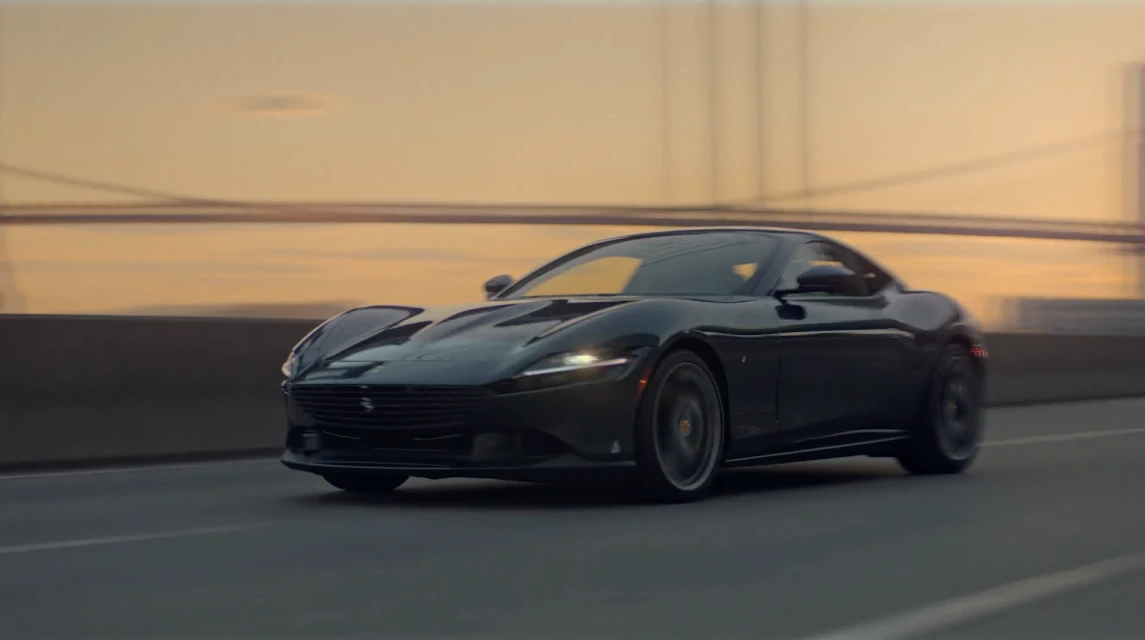 Sleek dark grey Ferrari Roma coupe speeding on a highway with blurred motion against a golden sunset background and bridge structure.