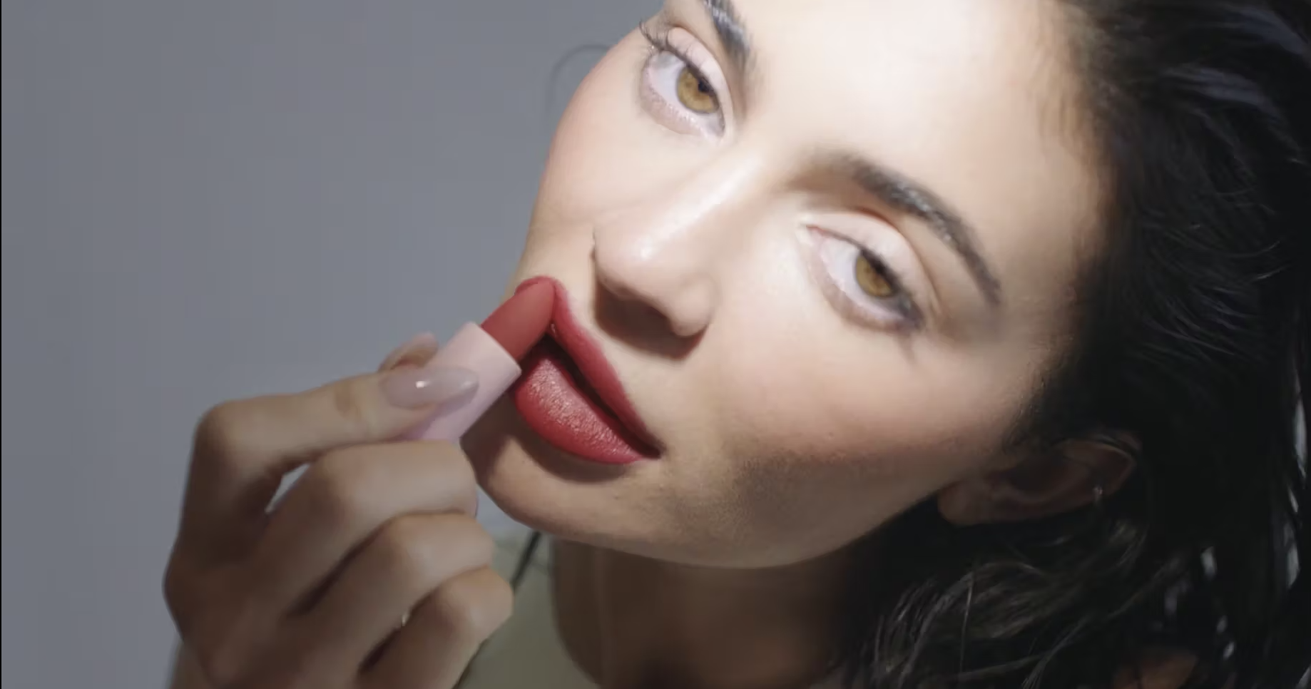 Close-up shot of an adult woman applying brick-red lipstick, focusing on her face, hand, and the cosmetic product in a beauty context.