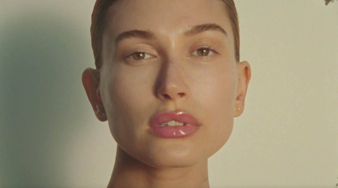 Close-up portrait of a young adult female face with fair skin, subtle make up, and glossy pink lips against a neutral background.