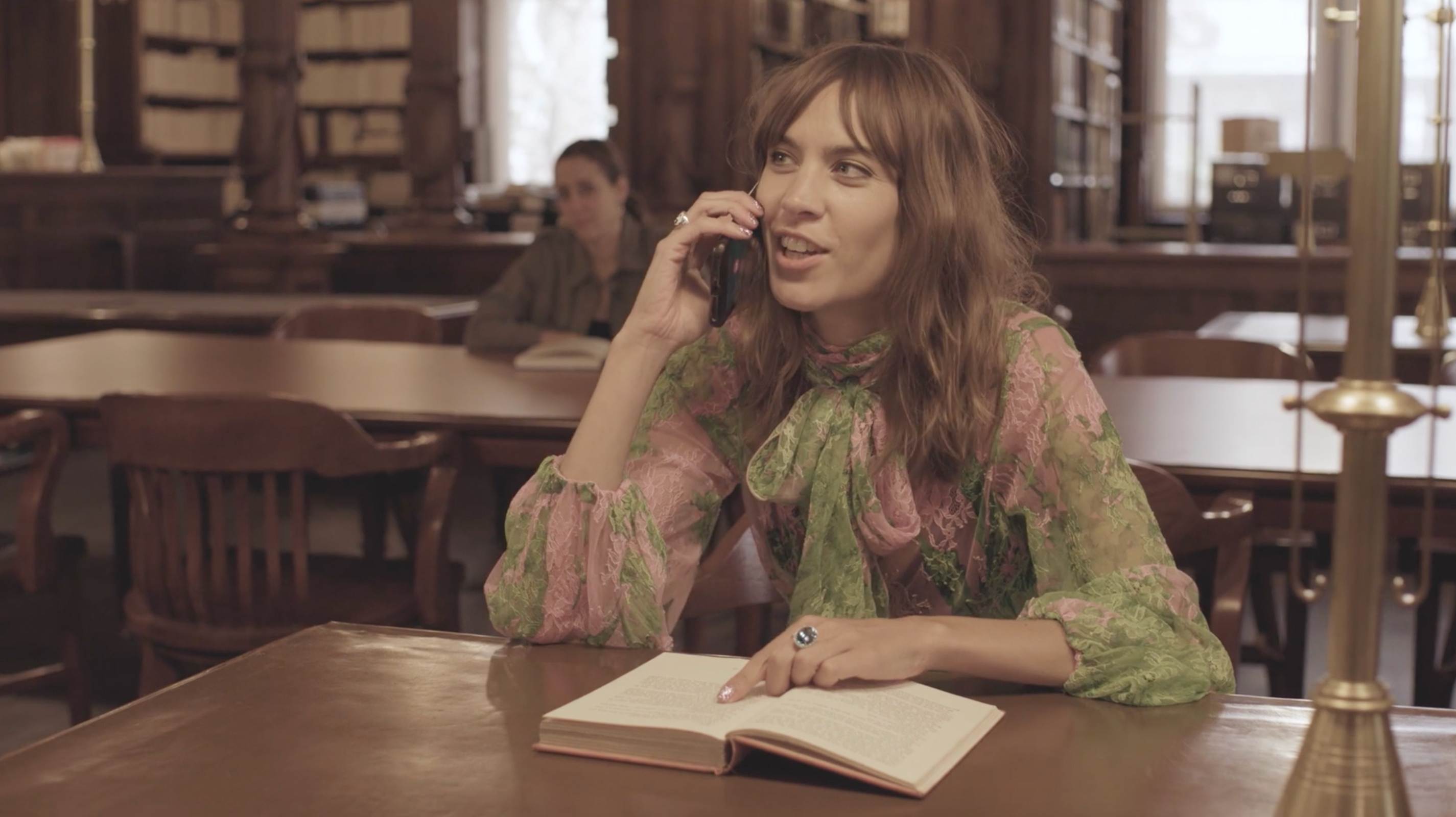 A woman with long brown hair sits at a table in a library, talking on a phone while pointing at an open book, with bookshelves behind her.