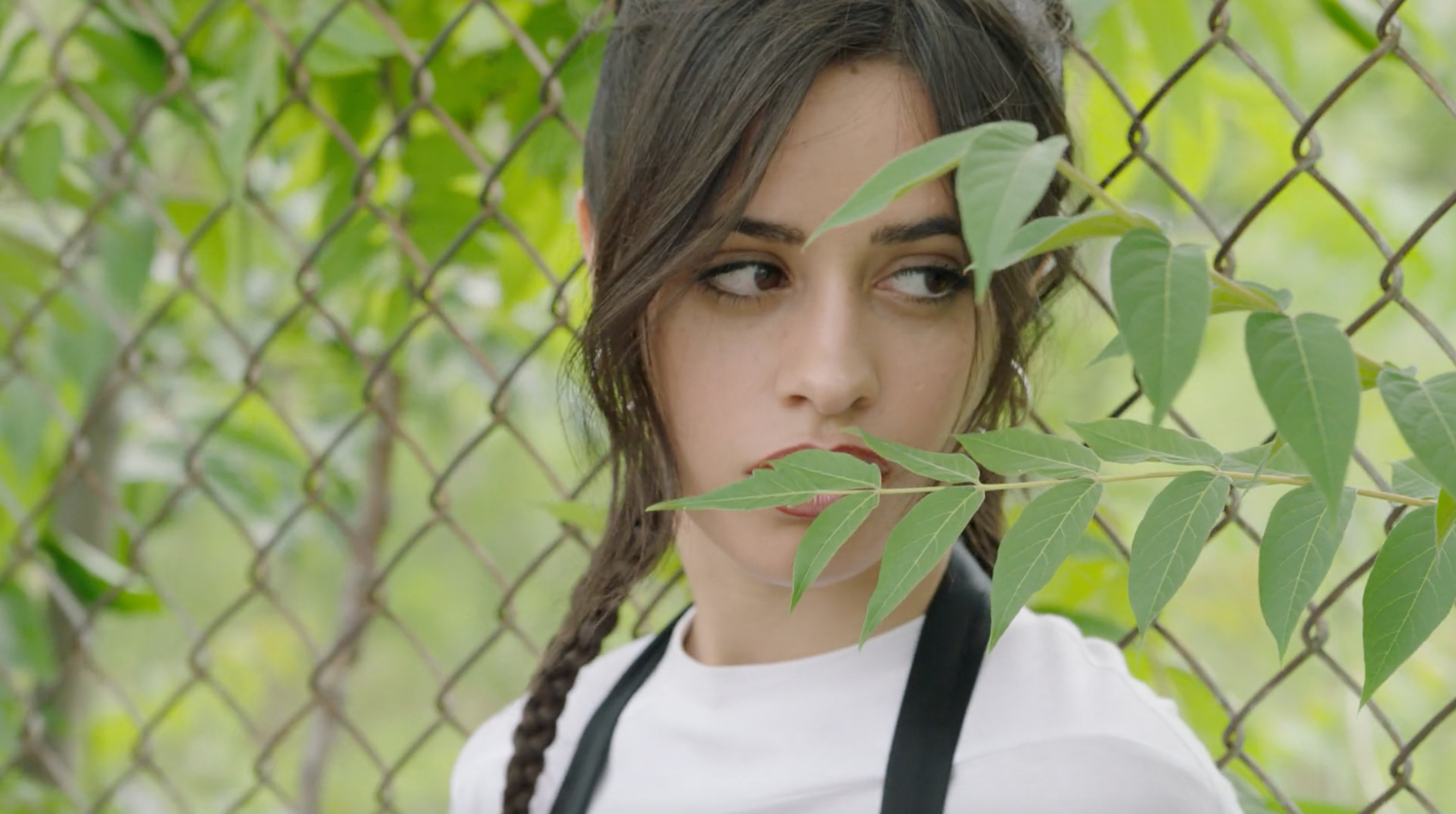 Close-up of a brunette woman with braided pigtails standing near a chain link fence outdoors with green foliage in the foreground.