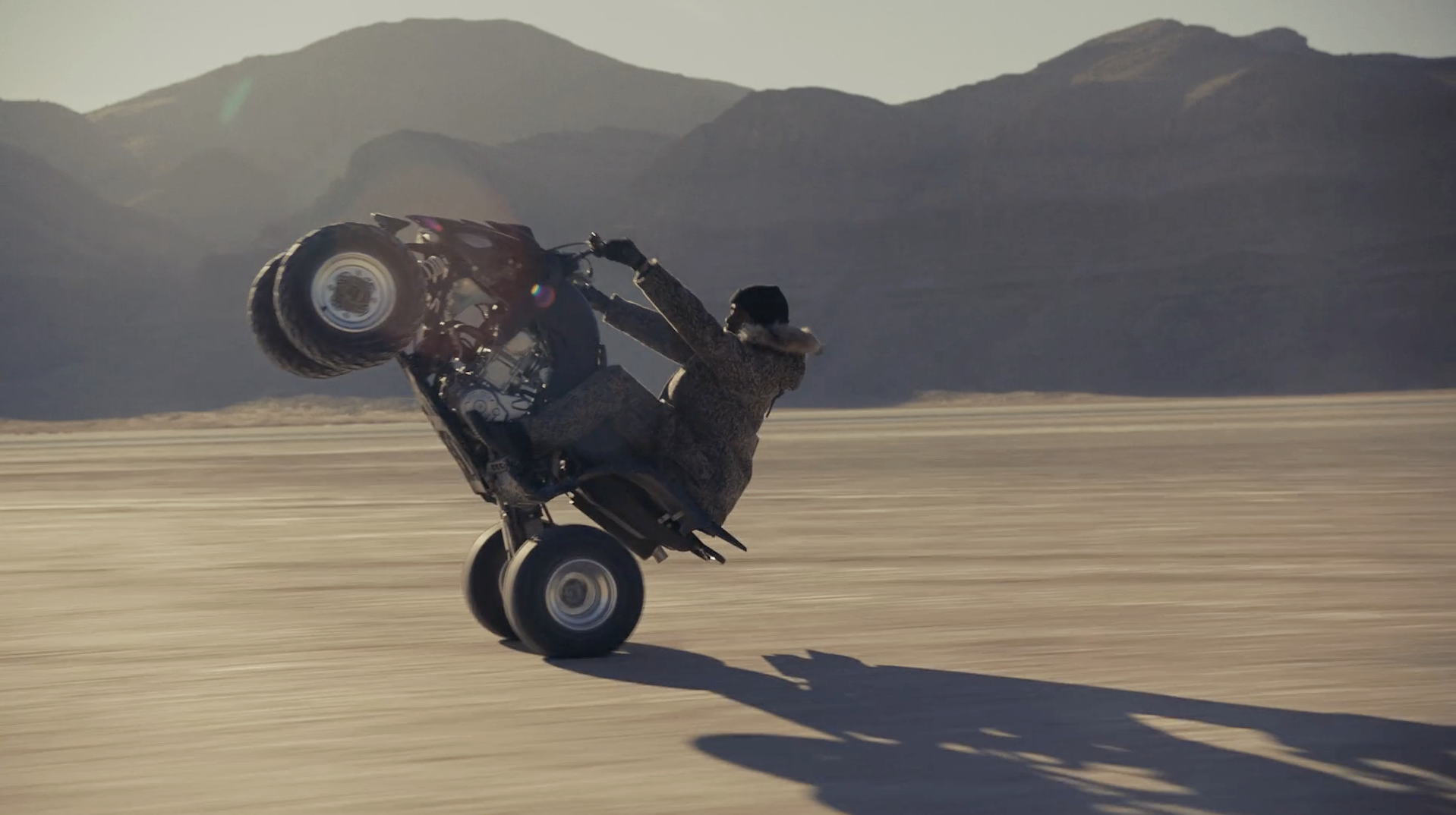 An adult rides an ATV doing a wheelie on a desert landscape with distant mountains under a clear sky in the daytime.