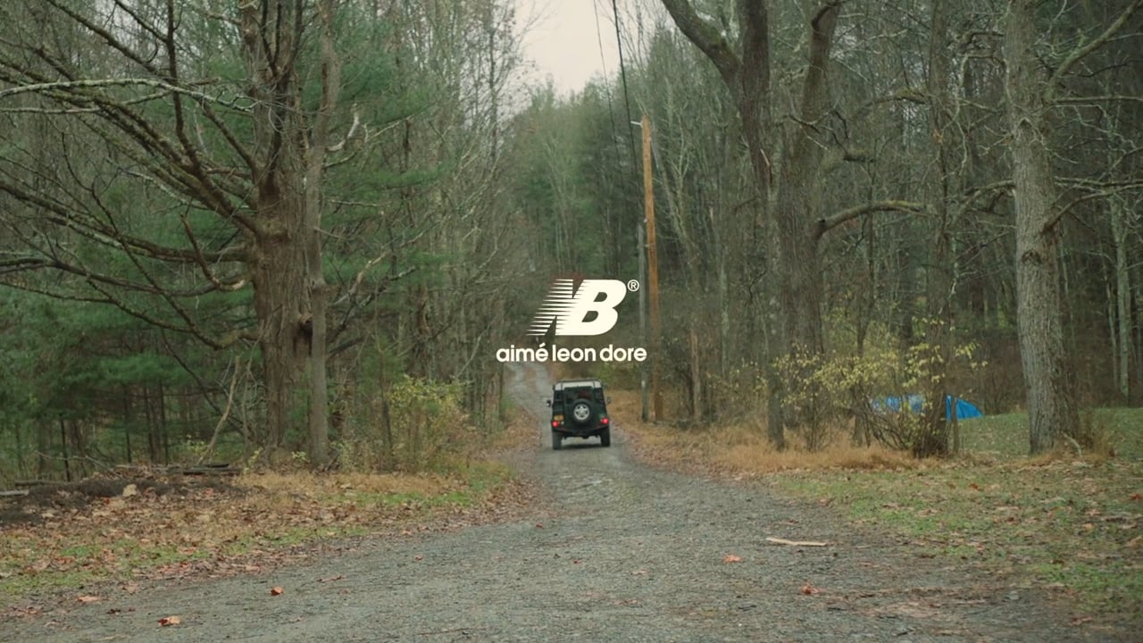 SUV drives down tree-lined gravel road under fashion brand logo. New Balance and Aime Leon Dore, countryside, green and brown tones.