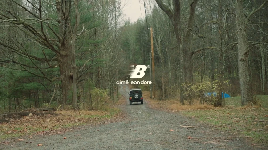 SUV drives down tree-lined gravel road under fashion brand logo. New Balance and Aime Leon Dore, countryside, green and brown tones.