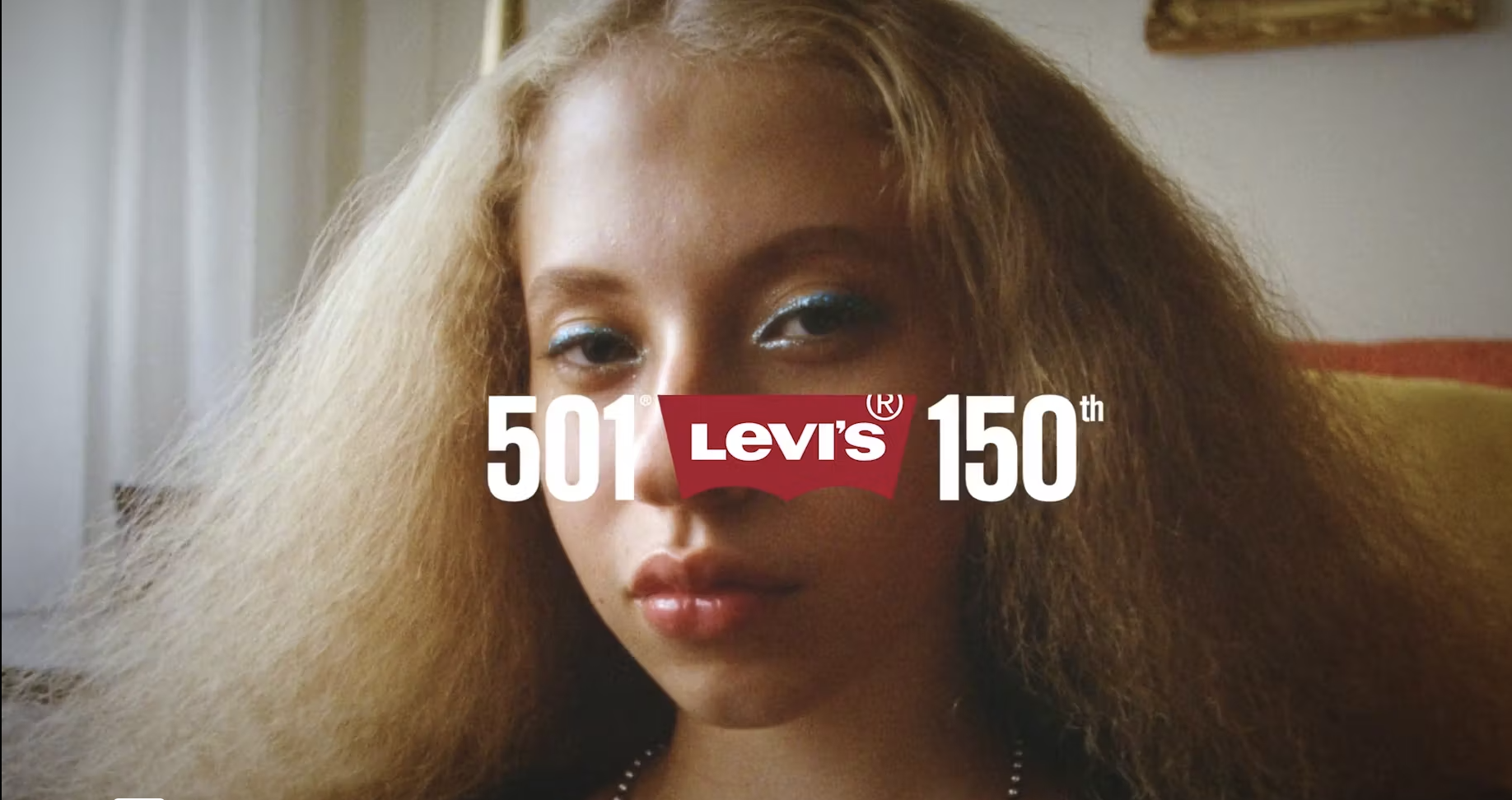 Close-up shot of an adult woman with blonde hair and blue eyeshadow in front of a Levi's 501 150th anniversary logo in an indoor setting.