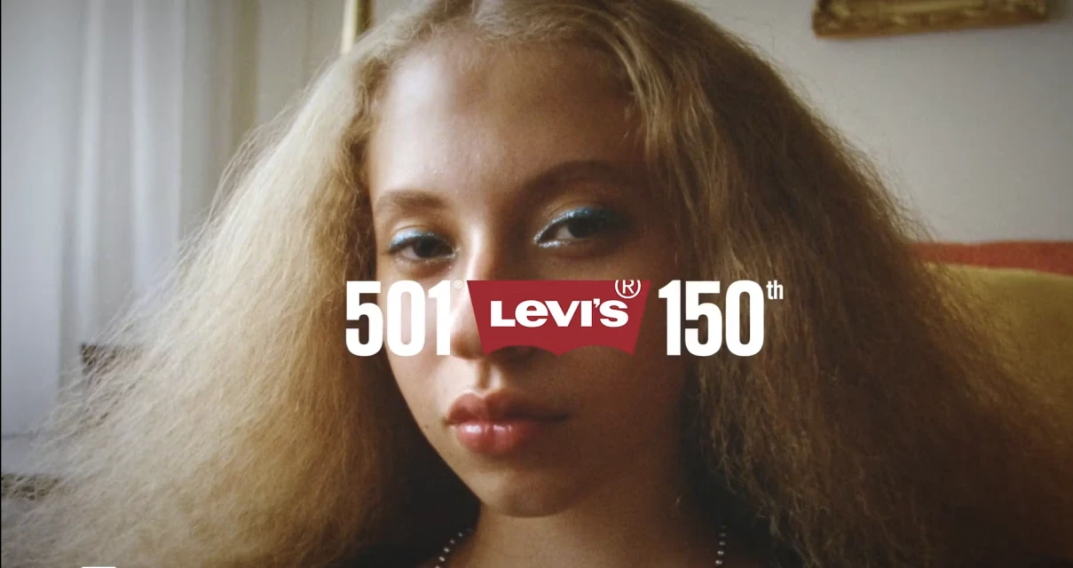 Close-up shot of an adult woman with blonde hair and blue eyeshadow in front of a Levi's 501 150th anniversary logo in an indoor setting.