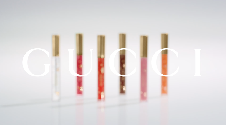 Six Gucci Rouge De Beauté Brillant lipsticks in varying colors stand on a white surface with the brand's logo superimposed in white.