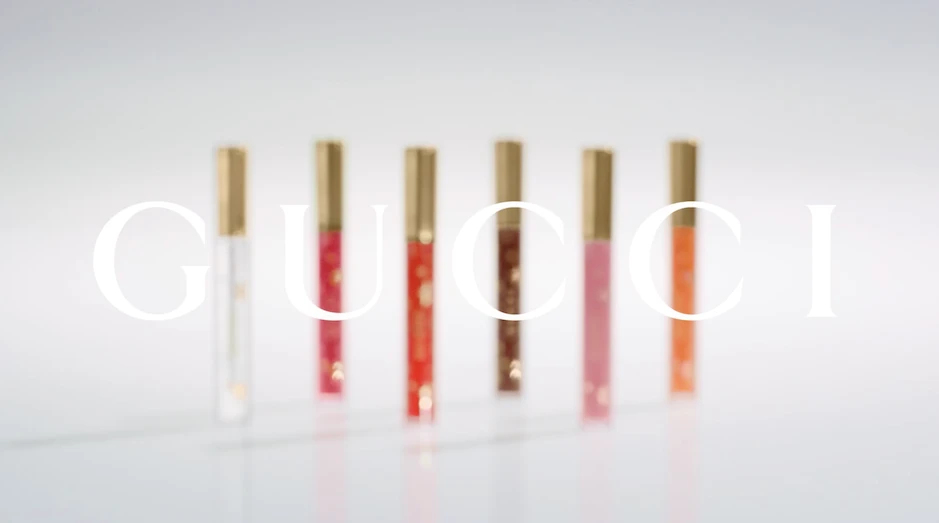 Six Gucci Rouge De Beauté Brillant lipsticks in varying colors stand on a white surface with the brand's logo superimposed in white.