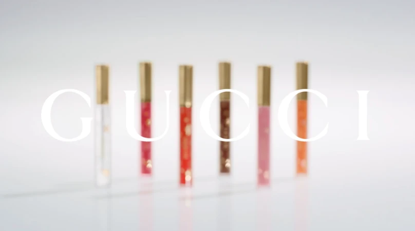Six Gucci Rouge De Beauté Brillant lipsticks in varying colors stand on a white surface with the brand's logo superimposed in white.