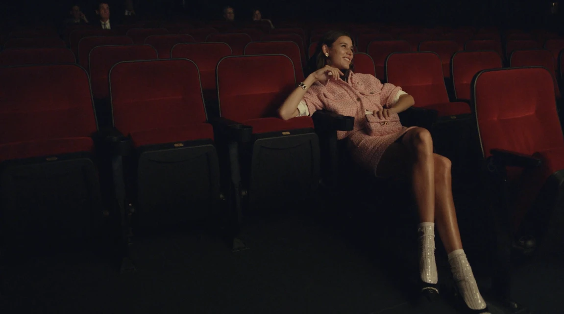 A woman in a pink skirt suit is seated in a red theater seat, smiling slightly with her legs extended and some other people behind her.