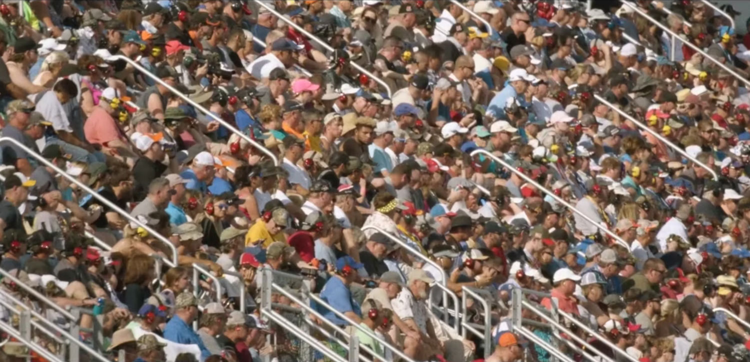 Large crowd of spectators fill stadium seating, enjoying an outdoor event with a mix of hats, shirts, and sunlit views, creating a lively atmosphere.